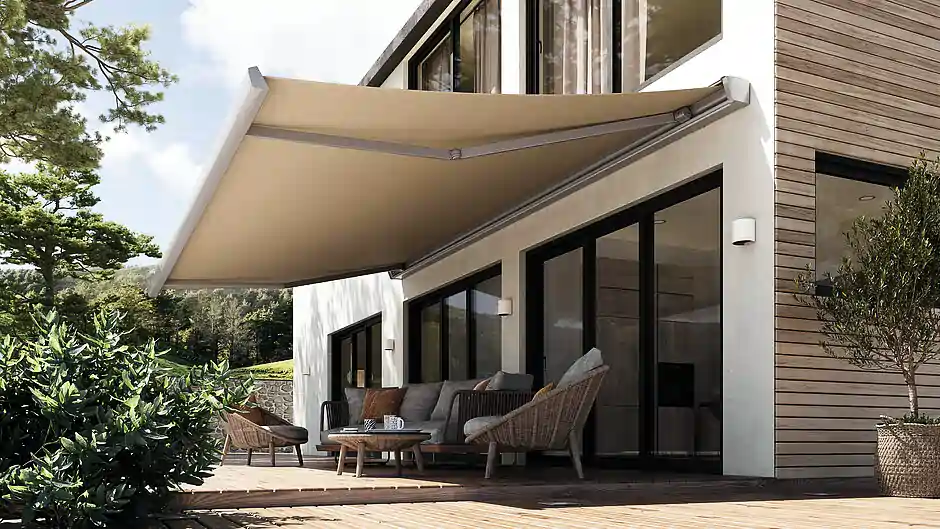 An der Fassade eines modernen Hauses mit großen Fenstern und einer Holzverkleidung ist eine beige Markise ausgefahren. Unter der Markise steht eine Sitzgruppe mit einem Sofa und zwei Sesseln aus geflochtenem Material sowie einem niedrigen Tisch. Rechts neben der Sitzgruppe steht ein großer Blumentopf mit einem kleinen Baum. Im Vordergrund sind grüne Pflanzen zu sehen, und im Hintergrund ragen Bäume in den Himmel.