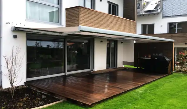 Eine weiße Markise ist an der Fassade eines Hauses mit Holzverkleidung und weißem Putz montiert. Die Markise ist ausgefahren und spendet Schatten auf einer Holzterrasse. Auf der Terrasse steht ein mit schwarzer Abdeckung geschützter Grill sowie zwei dunkelgraue Stühle mit einem kleinen Tisch dazwischen. Im Hintergrund sind Bäume und eine grüne Wiese zu sehen. Ein Glasgeländer begrenzt die Terrasse nach außen hin.