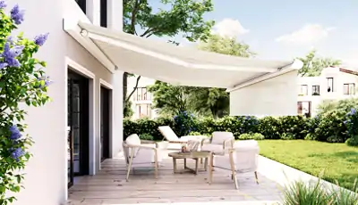 Eine weiße, ausfahrbare Kasettenmarkise der Marke MHZ spannt sich über eine helle Holzterrasse an einem modernen Haus. Unter der Markise stehen ein kleiner Tisch und mehrere Stühle mit hellen Polstern. Im Hintergrund ist ein gepflegter Garten mit grünem Rasen, Hecken und Bäumen zu sehen. Die Hausfassade ist hell und hat große Fenster, die zur Terrasse führen. Der Himmel ist blau mit einigen weißen Wolken.