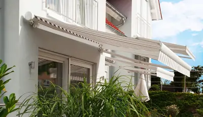 An der Fassade eines weißen Hauses sind mehrere ausfahrbare Hülsenmarkise mit einem gestreiften Muster in Beige- und Brauntönen angebracht. Die Markisen sind teilweise ausgefahren und spenden Schatten über den Fenstern und einer Terrasse. Unter den Markisen befinden sich große Fenster und eine Tür. Im Vordergrund wachsen grüne Pflanzen, und im Hintergrund ist ein blauer Himmel mit einigen Wolken zu sehen.