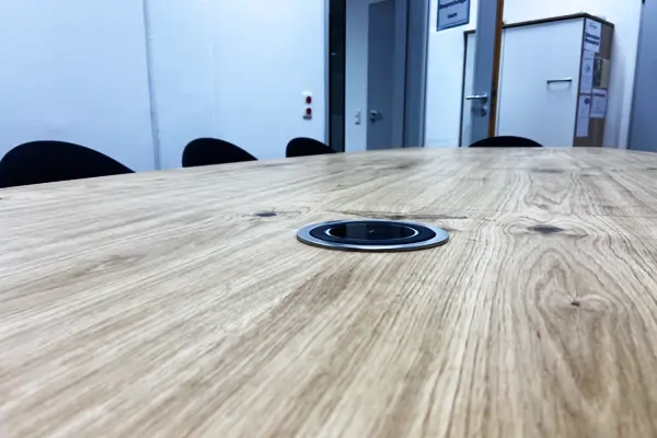 Ein Konferenztisch mit einer sichtbaren Holzstruktur aus Eiche, die Maserung und Knoten zeigt. In der Mitte des Tisches befindet sich eine runde, schwarze Kabeldurchführung. Im Hintergrund sind schwarze Stühle und ein weißer Raum mit Türen und einem Schrank zu sehen.