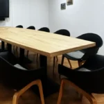 Ein rechteckiger Konferenztisch aus hellem Multiplex-Eichenholz steht in einem weißen Raum. Um den Tisch herum sind schwarze Stühle mit hellen Holzbeinen angeordnet. An der Wand im Hintergrund hängt ein großer Flachbildschirm. Der Boden besteht aus Holzlaminat in einem warmen Braunton.