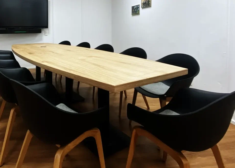 Ein rechteckiger Konferenztisch aus hellem Multiplex-Eichenholz steht in einem weißen Raum. Um den Tisch herum sind schwarze Stühle mit hellen Holzbeinen angeordnet. An der Wand im Hintergrund hängt ein großer Flachbildschirm. Der Boden besteht aus Holzlaminat in einem warmen Braunton.