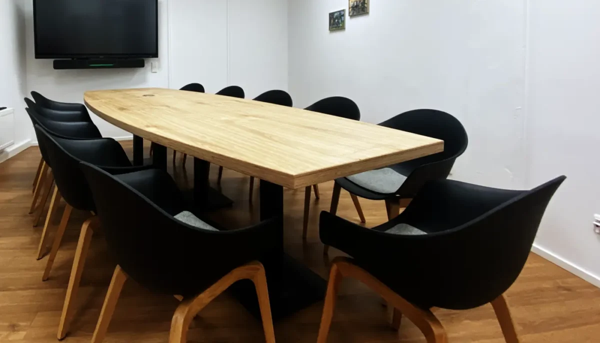 Ein rechteckiger Konferenztisch aus hellem Multiplex-Eichenholz steht in einem weißen Raum. Um den Tisch herum sind schwarze Stühle mit hellen Holzbeinen angeordnet. An der Wand im Hintergrund hängt ein großer Flachbildschirm. Der Boden besteht aus Holzlaminat in einem warmen Braunton.