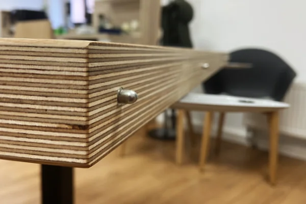 Nahaufnahme einer Kante eines mehrschichtigen Multiplex-Tischplatte mit einem Metallverbinder, unscharfer Hintergrund mit Stuhl und Tisch.