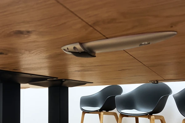 Unterseite eines Holztisches in Eiche mit Metallhalterung als verbinder der Tischplatte, dahinter drei schwarze Stühle mit Holzbeinen.